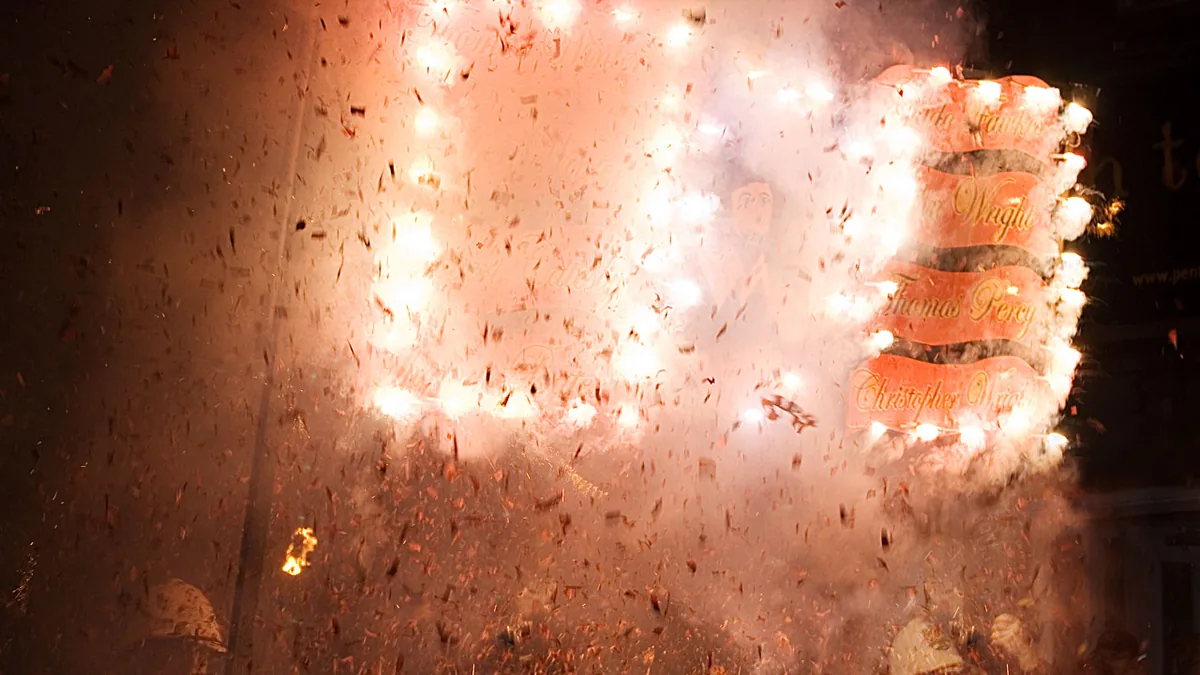 Lewes Borough Bonfire Society members in colonial soldiers' white helmets carrying a Guy Fawkes banner as the bangers covering it explode in sparks and smoke