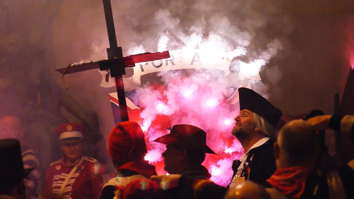Pink flares lighting the smoke around costumed society members and a tall cross on Bonfire Night