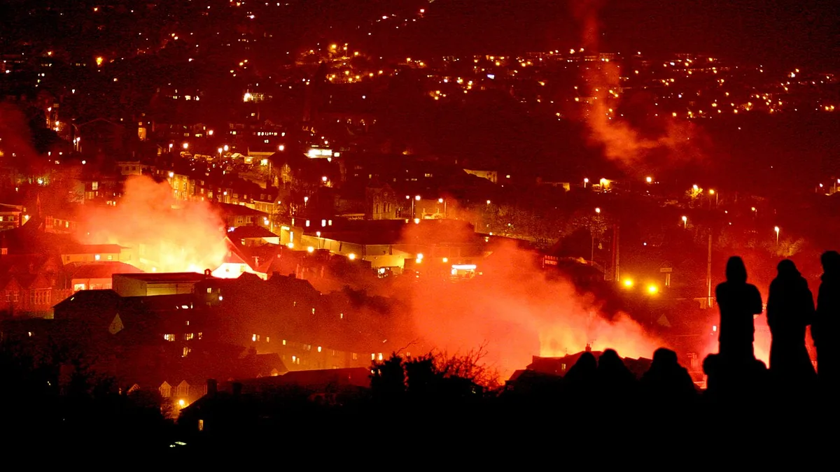 Watchers silhouetted on a hillside above Lewes, with firelight and red smoke rising over the town at night